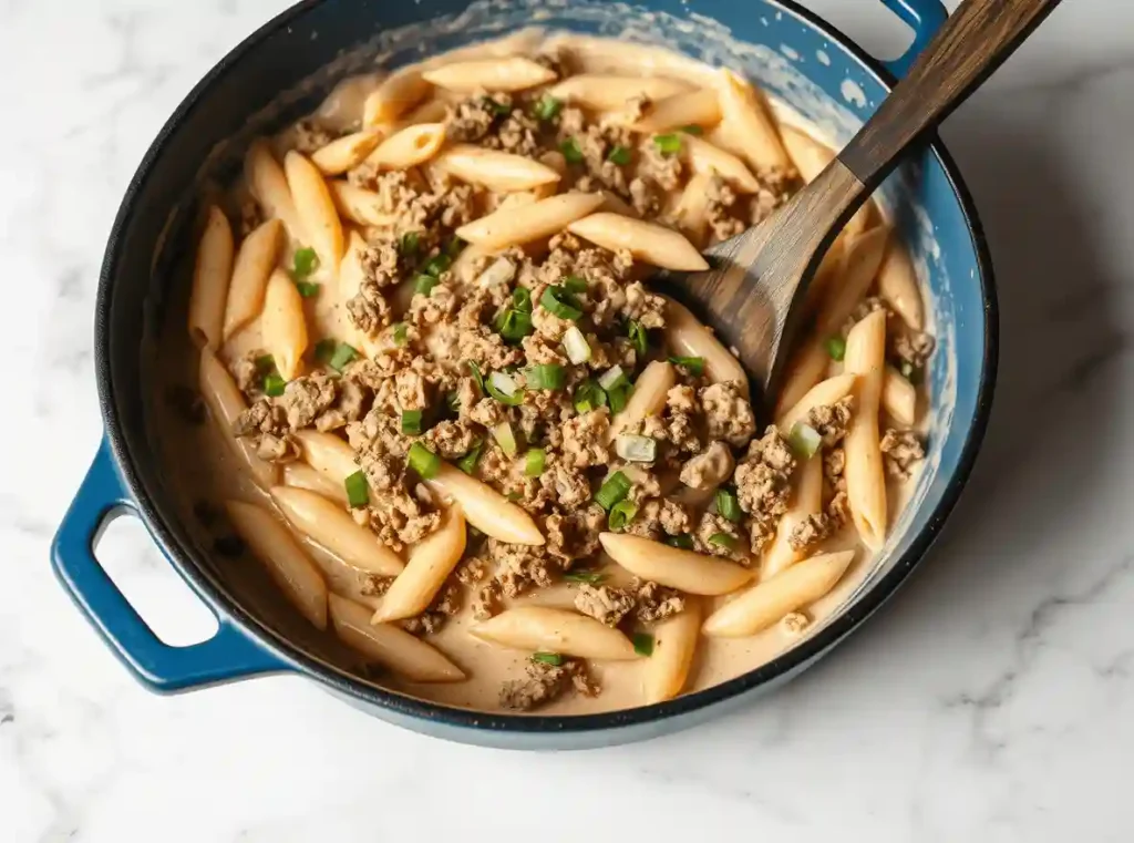 Large cast-iron skillet filled with creamy sausage pasta, featuring penne noodles in a velvety white sauce, garnished with sautéed spinach and sliced Italian sausage.