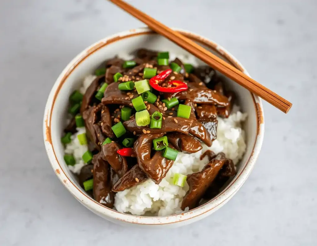 Mongolian Beef in a white ceramic bowl with a brown rim, topped with green scallions, sesame seeds, and red chili peppers. Served with wooden chopsticks on a neutral gray background.