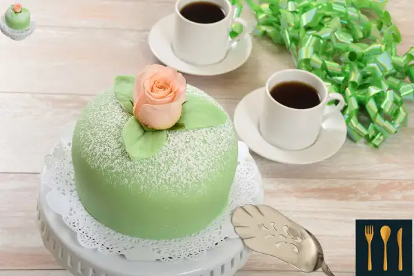 A Swedish Princess Cake topped with a rose, served on a white cake stand next to two cups of black coffee and a green gift ribbon.