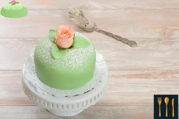A whole Swedish Princess Cake topped with a pink rose and powdered sugar, displayed on a white cake stand with a lace doily.